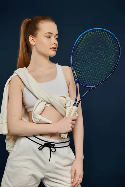 Una joven en ropa deportiva posa con una raqueta de tenis sobre un vibrante telón de fondo azul. - foto de stock