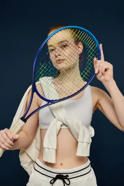 Una giovane donna tiene una racchetta da tennis davanti al viso, posando su un vibrante sfondo blu. — Foto stock