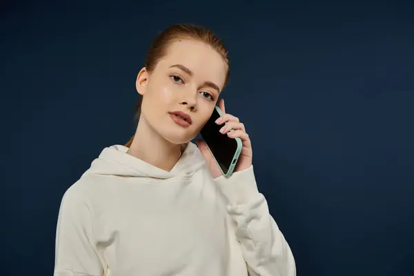 A young woman in a white hoodie makes a phone call against a deep blue backdrop. — Stock Photo