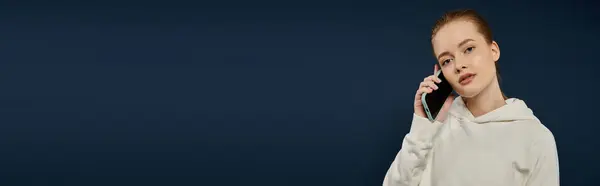 A young woman in a white hoodie talks on her phone against a blue backdrop. — Stock Photo