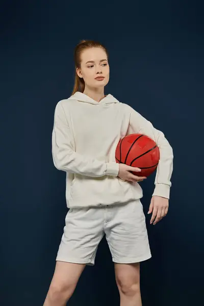 Eine junge Frau in weißen Shorts und Kapuzenpulli posiert mit einem Basketball vor blauem Hintergrund. — Stockfoto