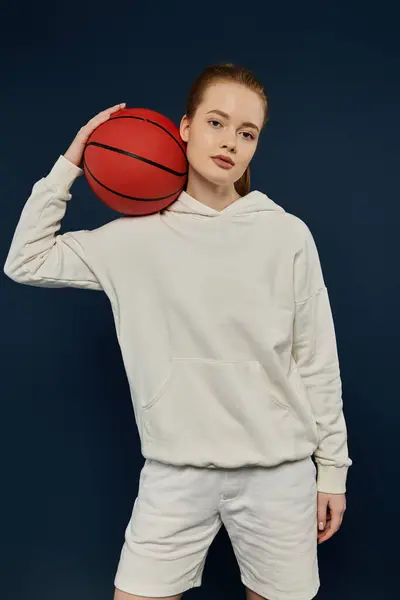 A young woman in a white hoodie and shorts poses with a basketball against a vibrant blue backdrop. — Stock Photo