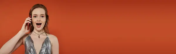A young woman with a surprised expression speaks on a phone against a vibrant orange backdrop. — Stock Photo