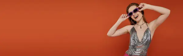 Woman smiles brightly in silver top and sunglasses against orange background. — Stock Photo