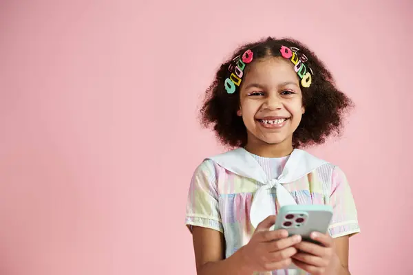 A young African American girl with colorful hair clips smiles while holding a phone against a pink background. — Stock Photo