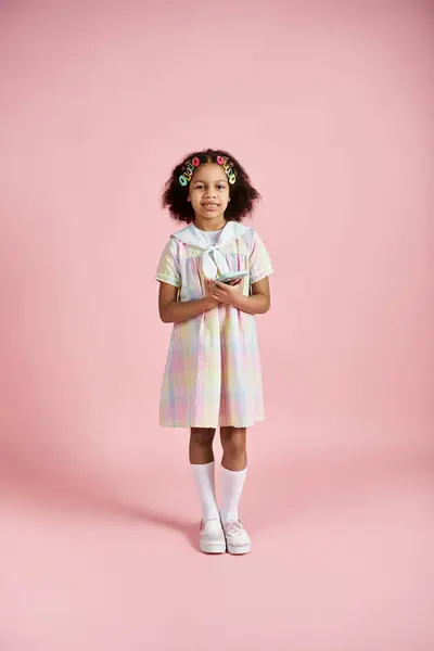 A young African American girl in a pastel dress with hair clips stands on a pink background. — Stock Photo