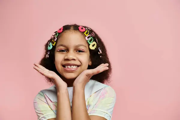 Une jeune afro-américaine avec des pinces à cheveux colorées sourit vivement dans une robe rose sur un fond rose. — Photo de stock