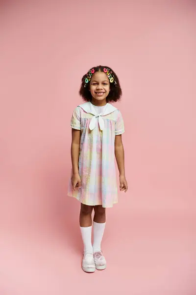 A young African American girl with colorful hair clips stands in a pastel dress, smiling against a pink backdrop. — Stock Photo