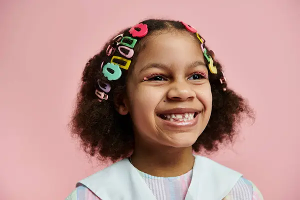 Une jeune fille afro-américaine avec des pinces à cheveux colorées sourit vivement sur un fond rose. — Photo de stock