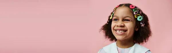 A young African American girl with colorful hair clips smiles brightly against a pink background. — Stock Photo