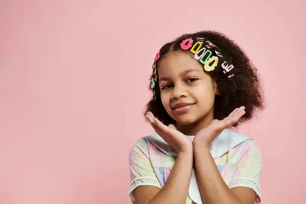 Young Black girl with cute hairstyle and colorful hair clips poses in pastel dress on pink background. — Stock Photo