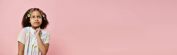 Uma garota afro-americana fofa com grampos de cabelo coloridos fica contra um fundo rosa, olhando atentamente para cima. — Fotografia de Stock