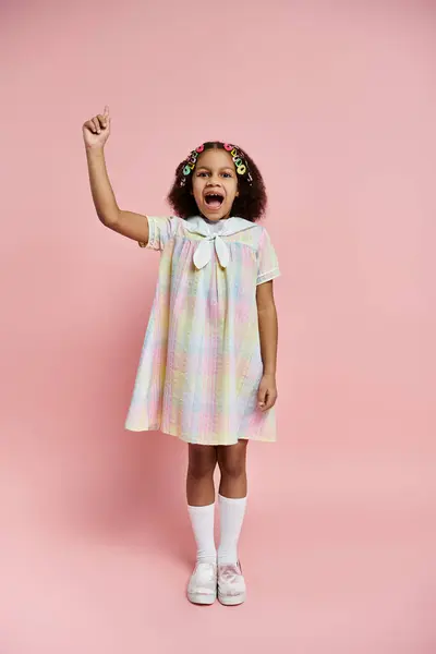 A young African American girl with bright hair clips stands in a colorful dress, pointing upwards on a pink background. — Stock Photo