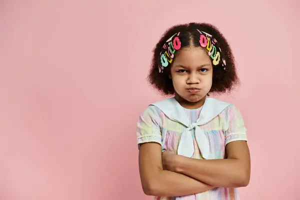 An African American girl with colorful hair clips stands with crossed arms in a pink and white dress on a pink background. — Stock Photo