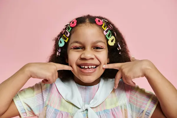 Une jeune afro-américaine dans une robe colorée sourit vivement avec ses doigts sur les joues, sur fond rose. — Photo de stock
