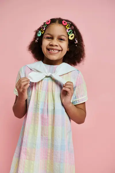 Une jeune afro-américaine avec des pinces à cheveux colorées sourit vivement dans une robe colorée, debout sur un fond rose. — Photo de stock