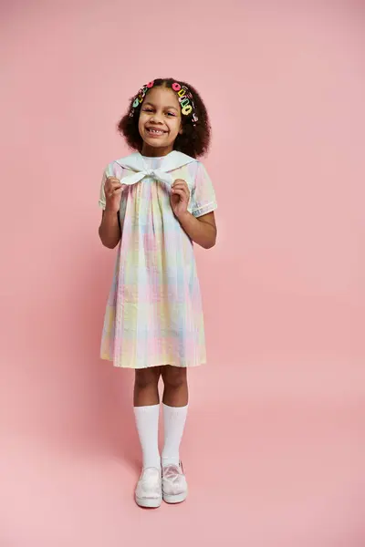 Симпатичная афроамериканка с заколками для волос ярко улыбается, стоя в радужном клетчатом платье на розовом фоне. — стоковое фото