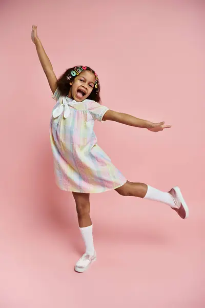 A young African American girl in a colorful dress and hair clips stands on a pink background, striking a playful pose. — Stock Photo