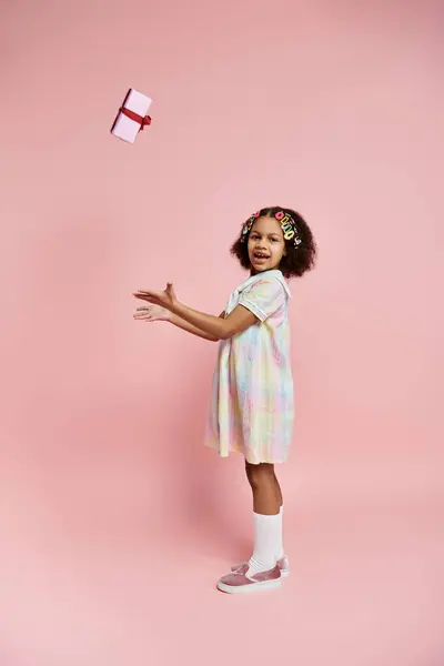 An African American girl in a colorful dress stands on a pink background, smiling as she tosses a gift box into the air. — Stock Photo