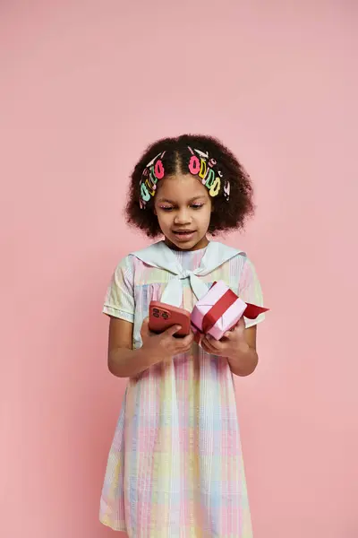 A young African American girl in a plaid dress, with colorful hair clips, holds a gift box and a smartphone against a pink background. — Stock Photo