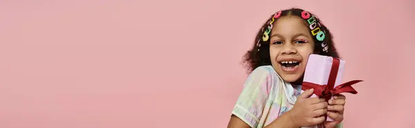 A young African American girl with colorful hair clips smiles brightly as she holds a wrapped gift in front of a pink background. — Stock Photo