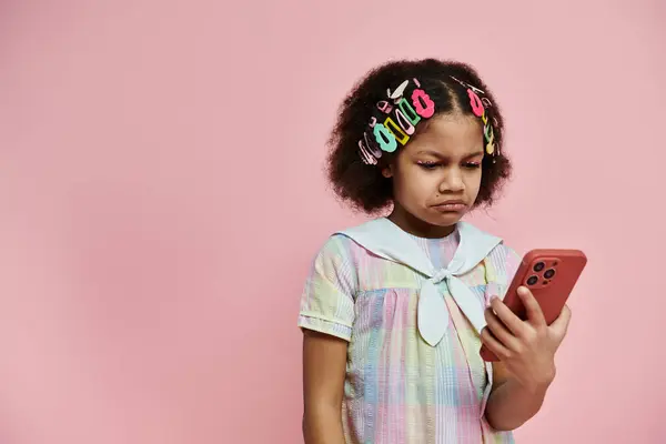 Uma menina afro-americana bonito com clipes de cabelo coloridos fica em um vestido listrado rosa e branco contra um fundo rosa. — Fotografia de Stock
