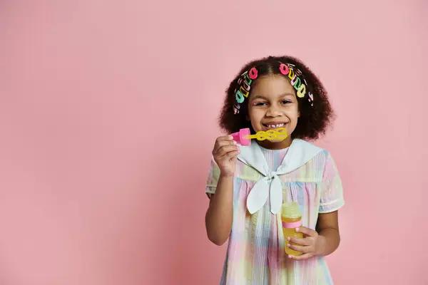 Young girl with colorful hair clips smiles brightly while holding a bubble wand and bottle of bubbles. She wears a cute, colorful dress. — Stock Photo