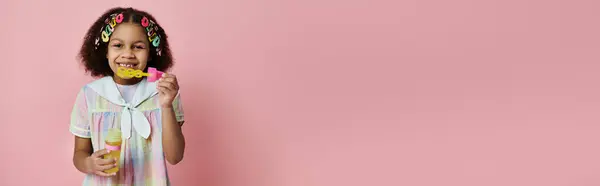 Una giovane ragazza afroamericana in un abito colorato si erge su uno sfondo rosa, soffiando bolle e sorridendo. — Foto stock