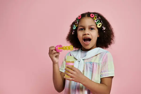 A young African American girl blows bubbles with a playful expression, wearing a colorful dress and hair clips. — Stock Photo