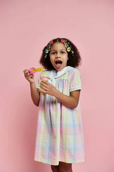 Una giovane ragazza afroamericana con clip di capelli colorati si erge in un abito rosa su uno sfondo rosa, soffiando bolle con una bacchetta. — Foto stock