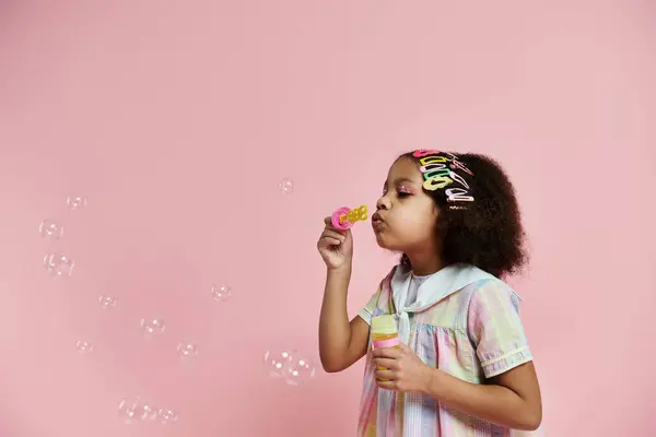 A young African American girl in a pink and white dress blows bubbles with a pink wand against a pink background. — Stock Photo