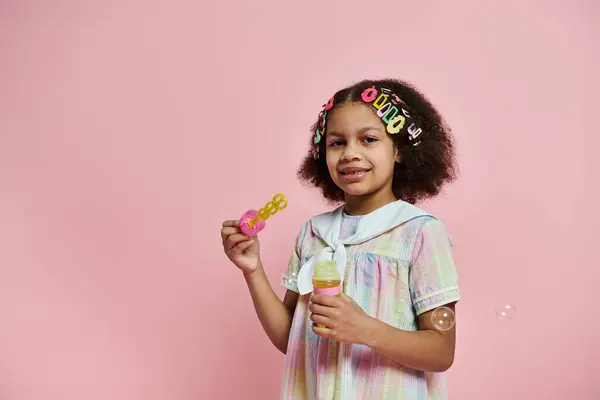 A young African American girl with colorful hair clips blows bubbles in a pink and white dress against a pink background. — Stock Photo