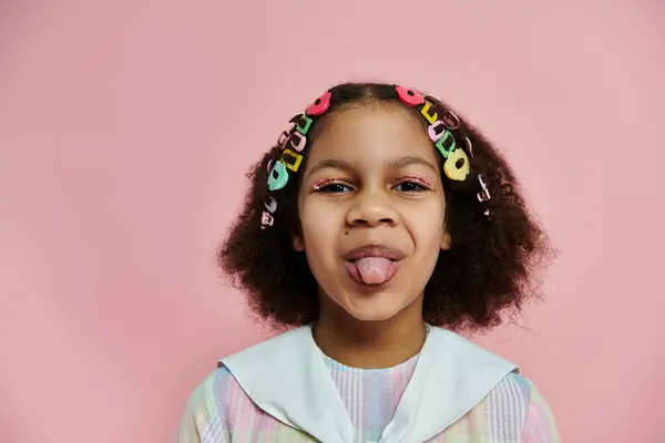 Une fille noire avec des pinces à cheveux colorées sort sa langue et sourit ludique devant un fond rose. — Photo de stock