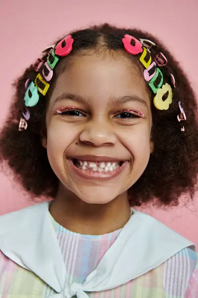 A young African American girl with curly hair adorned with colorful hair clips smiles brightly against a pink background. — Stock Photo