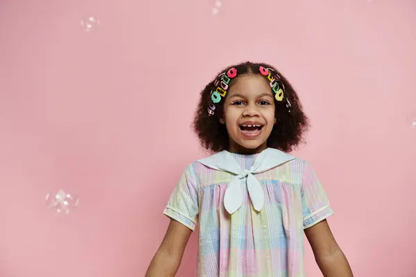 Young African American girl with hair clips in her hair smiles brightly in a pastel dress against a pink background. — Stock Photo