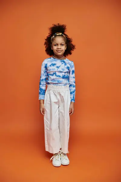 A young African American girl stands confidently in a trendy outfit with hair clips, on a bright orange background. — Stock Photo