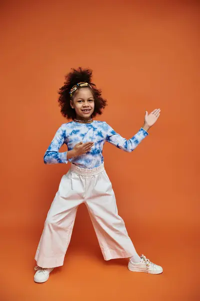 An African American girl with hair clips stands in a trendy outfit against an orange background, showcasing a cool and modern style. — Stock Photo