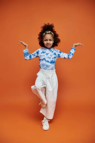 A young African American girl with a trendy hairstyle and outfit poses with her arms out in front of an orange backdrop. — Stock Photo