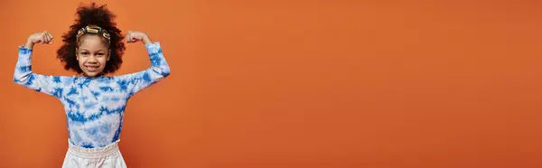 A young African American girl with hair clips stands confidently against an orange background, showcasing a trendy outfit. — Stock Photo