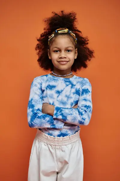 A young African American girl with a trendy tie-dye shirt and hair clips stands confidently against an orange backdrop. — Stock Photo