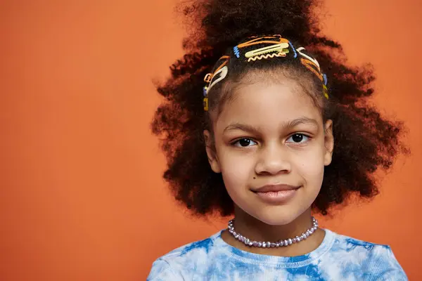 A young African American girl with colorful hair clips stands in a trendy outfit against a bright orange background. — Stock Photo