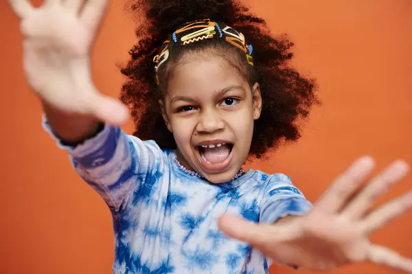 A young African American girl in a blue tie-dye shirt poses with her hands outstretched against an orange backdrop. — Stock Photo