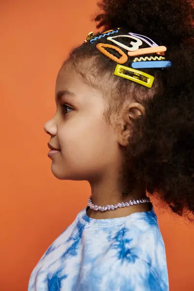 A chic Black girl with colorful hair clips stands on an orange background in trendy clothes with a beautiful hairstyle. — Stock Photo