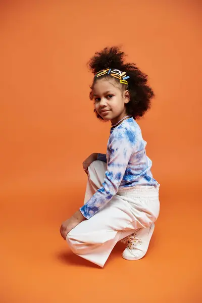 An African American girl in a stylish tie-dye shirt and white pants poses against a vibrant orange backdrop. — Stock Photo