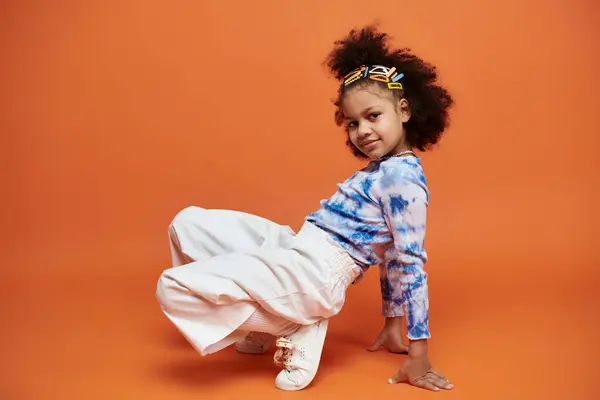 Colorful hair clips and trendy outfit on African American girl against orange background. Blue and white tie-dye shirt with white pants. — Stock Photo