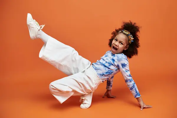 Stylish African American girl with hair clips standing in trendy blue and white shirt and white pants on orange background. — Stock Photo