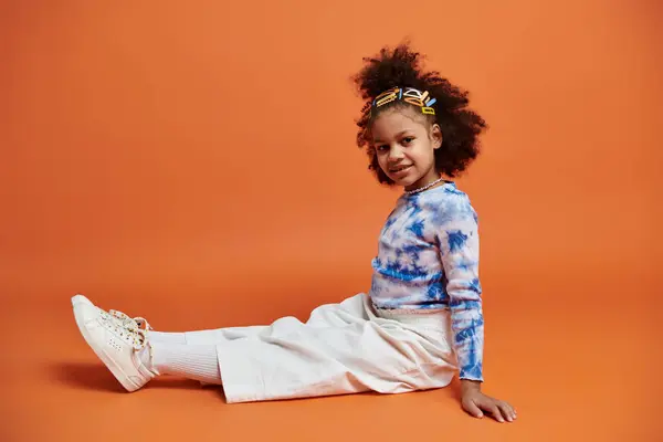 A young African American girl with colorful hair clips sits on an orange background wearing a tie-dye shirt and white pants. — Stock Photo