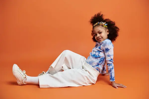 A young African American girl wearing a tie-dye shirt, white pants, and hair clips poses on an orange background. — Stock Photo