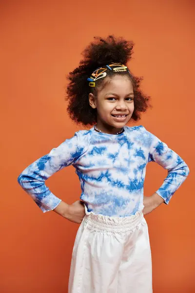 A young African American girl with colorful hair clips stands confidently against an orange background, wearing a trendy tie-dye shirt. — Stock Photo