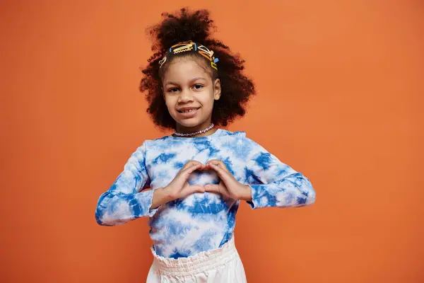 A young African American girl in a trendy outfit stands against a vibrant orange backdrop, smiling and making a heart shape with her hands. — Stock Photo